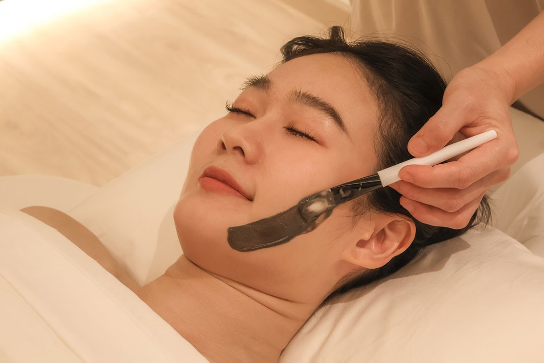 A woman receiving a facial treatment with a black face mask at Makkha Spa in Chiangmai, showcasing relaxation and professional skincare services.
