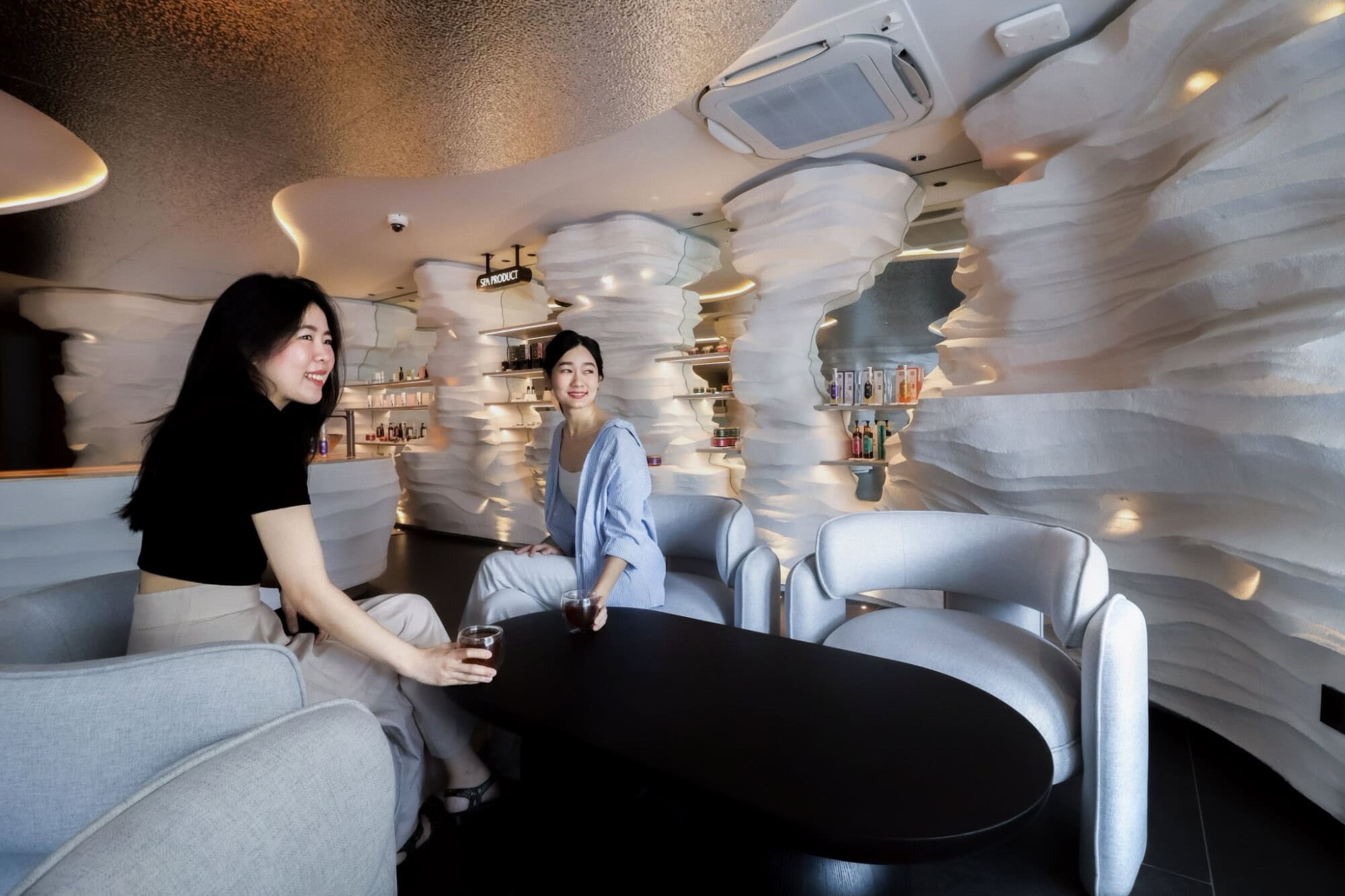 Relaxing spa lounge in Chiangmai with modern white textured walls, comfortable seating, and two women enjoying drinks, creating a serene environment at Makkha Spa.