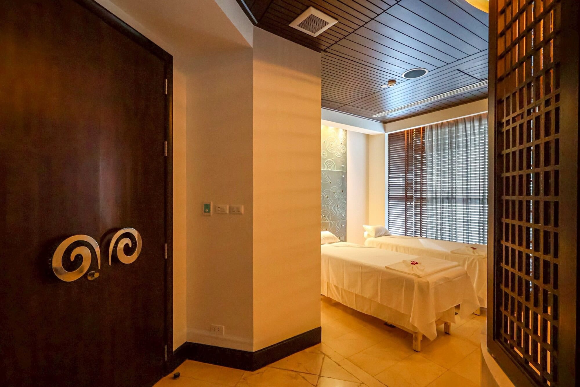 Relaxing spa treatment room with two massage beds, soft lighting, elegant decor, and large windows with wooden blinds in Chiang Mai at Makkha Spa – the best spa in Chiang Mai.